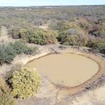 Aerial view of a small, muddy pond surrounded by sparse trees and dry grassland in a rural landscape under a clear sky.