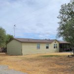 A beige manufactured home with a brown roof sits on a dry, grassy lot with scattered trees. A covered driveway with two vehicles is visible on the right, and the sky is partly cloudy.