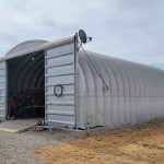 A large, silver metal Quonset hut stands on dry, rocky ground with its front doors open, revealing tools inside. A green tractor is parked nearby, and desert vegetation surrounds the building under a cloudy sky.
