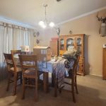A dining room with a wooden table and chairs, boxes and items on the table, a china cabinet, wall-mounted deer heads, and a gun safe. Light streams in through curtained windows.