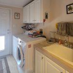 A laundry room with a washer and dryer side by side, stacked towels, a utility sink, overhead cabinets, and wall-mounted towels. Sunlight streams through a door with a frosted window.