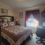 A cozy bedroom with a quilted bed, wooden furniture, a desk with a computer, shelves with books, an armchair, and a window with red curtains letting in natural light.