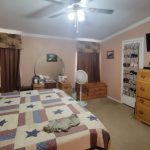 A cozy bedroom with a patriotic star-patterned quilt on the bed, wooden dressers, a TV mounted on the wall, a round mirror above a vanity, a standing fan, and a shoe organizer hanging on the door.