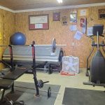 A home gym with a bench press, weights, a blue exercise ball, an elliptical machine, a tanning bed, a fan, and gym equipment. The walls are covered with plaques, awards, and framed pictures.