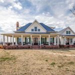 A large farmhouse with a wraparound porch, white siding, and a gray metal roof sits on a spacious, grassy lot under a partly cloudy sky. There are a few leafless trees and a stone walkway leading to the porch.
