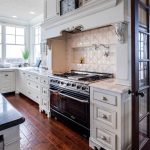 Bright, spacious kitchen with large windows, white cabinetry, a black countertop island with a sink, a stainless steel stove, and a glass door leading to a cozy dining area with a fireplace.