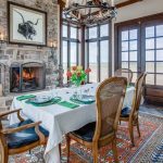 A dining room with a table set for six, white tablecloth, green napkins, and a vase of tulips. Large windows, a stone fireplace with a bull artwork, wooden chairs, and a chandelier decorate the space.