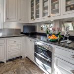 Bright kitchen with white cabinets, black countertops, stainless steel sink, small TV, bowl of oranges, orchids, and windows above the counter letting in natural light. The floor has large stone tiles.