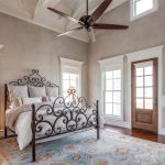 A bright, airy bedroom with a decorative wrought iron bed, plush bedding, wooden side table with a lamp, patterned armchair, large windows, a ceiling fan, and a blue patterned rug.