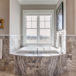 A luxurious bathroom with a large bathtub set in marble tile beneath a wide window, flanked by towels, artwork on the wall, and a wooden door to the right. Natural light fills the space.