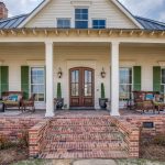 A large farmhouse with a wide, covered front porch featuring wicker furniture, green shutters, double wooden doors, and brick steps leading up to the entrance. The house has white siding and a metal roof.