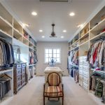 A spacious, organized walk-in closet with shelves and hanging clothes on both sides, dressers, a centered island, and a striped chair at the end. Natural light streams in from a window at the far end.