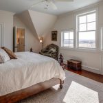 A bright bedroom with a large bed, wooden furniture, a chair by the window, and sunlight streaming through two windows, highlighting a neutral color scheme and hardwood floors.