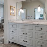 A bathroom with a marble-topped white vanity, large mirror, two wall-mounted lights, towel on a ring holder, and tile flooring. An adjacent room with a bathtub is visible in the background.