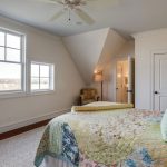 A bright bedroom with large windows, a ceiling fan, and a bed covered in colorful, patterned bedding. An upholstered chair sits in the corner near a door leading to an ensuite bathroom.