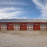 A red barn with a gray metal roof stands on a gravel lot under a partly cloudy sky. The barn has six doors with white X-shaped patterns and outdoor lights above each door. Open fields stretch into the distance.
