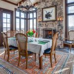A bright dining room with large windows, a stone fireplace, a table set for six with a white tablecloth, tulips centerpiece, wooden chairs, and a patterned rug on a tiled floor. A bull artwork hangs above the fireplace.