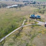 Aerial view of a rural property with a large house, red barn, winding driveway, scattered trees, fenced areas, and open fields stretching into the distance under a clear sky.