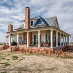 A charming farmhouse with a large wraparound porch, brick chimneys, and metal roof sits on a spacious, grassy property under a partly cloudy sky. There are rocking chairs on the porch and leafless trees nearby.