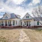 A large, white, two-story farmhouse with a covered front porch, green shutters, and a metal roof stands on a spacious, grassy lot under a partly cloudy sky. A stone path leads to the front steps.