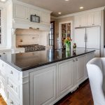 A spacious, elegant kitchen with white cabinets, a large black countertop island, stainless steel appliances, and wooden floors. Fresh flowers are on the island and a dining table with white chairs is visible in the foreground.