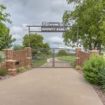 A gated entrance to "Divine Serenity Ranch," with brick pillars, metal gates, and lush landscaping, featuring green trees, shrubs, and a cloudy sky in the background.