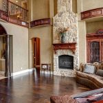 Spacious living room with dark wood floors, a stone fireplace, ornate furniture, and tall ceilings. A curved staircase and balcony are visible, with a large mantel and built-in shelving flanking the fireplace.