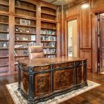A luxurious home office with rich wood paneling, built-in bookshelves, a large ornate desk, a leather chair, and polished wooden floors. An open doorway reveals a hallway with elegant decor.