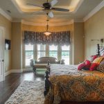 A spacious bedroom with a large ornate bed, patterned bedding, decorative pillows, a plush rug, dark wood floors, a ceiling fan, and a window nook with a green loveseat, framed by drapes and overlooking greenery.