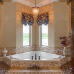 Luxurious bathroom with a white corner bathtub surrounded by mosaic tiles, flanked by two ornate columns. A chandelier hangs above, with decorative curtains on two windows and vases placed on ledges.