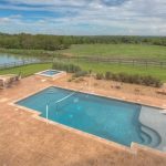 A backyard with a rectangular swimming pool, hot tub, and seating area is surrounded by a patio, grassy lawn, and a wooden fence overlooking a pond and open green fields under a partly cloudy sky.