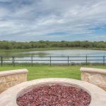 A stone fire pit filled with red rocks sits on a patio overlooking a grassy lawn, a wooden fence, and a small pond, with trees and a partly cloudy sky in the background.