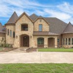 A large, two-story stone and brick house with steep, turret-like roofs and arched entryway, surrounded by a well-kept lawn and set against a blue sky with scattered clouds.