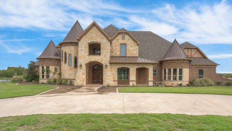 A large, two-story stone and brick house with steep, turret-like roofs and arched entryway, surrounded by a well-kept lawn and set against a blue sky with scattered clouds.