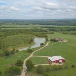 Aerial view of a rural landscape with green fields, scattered trees, a winding road, a pond, a red barn, and a house under a partly cloudy sky.