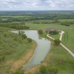 Aerial view of a rural landscape with a winding pond, a distant house surrounded by trees, and expansive green fields under a partly cloudy sky.