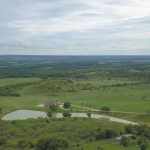 Aerial view of a rural landscape with green fields, scattered trees, a house near a pond, and a barn. The sky is overcast, creating a peaceful countryside scene stretching toward the horizon.
