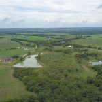 Aerial view of a rural landscape with green fields, scattered houses, winding roads, small ponds, and clusters of trees under a cloudy sky.