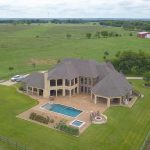 Aerial view of a large stone house with a spacious backyard, swimming pool, and hot tub, surrounded by green lawns and fencing, set in a rural landscape with fields and scattered trees.
