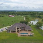 A large house with a pool sits on a spacious, fenced green property near a pond, surrounded by fields and trees. A winding driveway leads to a red barn in the distance. Multiple vehicles are parked near the house.