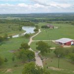 Aerial view of a rural landscape with a winding road, a red barn, scattered trees, green fields, and a small pond under a partly cloudy sky.
