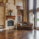 Spacious living room with high ceilings, large windows, a stone fireplace, a brown sofa, a grand piano, and wood floors. Natural light fills the room, and decorative cabinets and shelves add elegance.