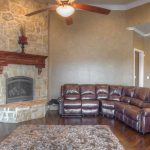 A cozy living room with a large brown leather sectional sofa, a stone fireplace with a wooden mantel, dark hardwood floors, and a ceiling fan with lights. A plush rug lies in front of the sofa.