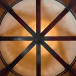 A circular wooden ceiling with dark wooden beams radiating from the center, creating a symmetrical, star-like pattern. Warm lighting highlights the wood tones and the smooth, beige surface between the beams.
