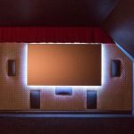 A home theater room with a large blank screen, red curtains at the top, four black speakers mounted on a patterned wall, and soft ambient lighting behind the screen.