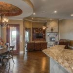 Spacious open-concept living area with a round dining table, decorative ceiling, marble kitchen counter, leather sofas, stone fireplace, wood floors, and large windows letting in natural light.