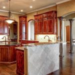 Spacious, elegant kitchen with dark wood cabinets, marble countertops, a central island, stainless steel appliances, and ornate columns leading to a living area with a stone fireplace and piano.