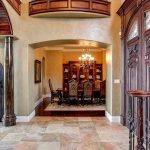 Elegant entryway with ornate wooden double doors, wrought iron details, marble tile floor, and an arched view into a dining room with a chandelier and tall-backed chairs around a dining table.