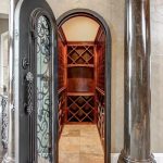 A small wine cellar with wooden racks and shelves is visible through an ornate iron and glass arched door, flanked by two dark marble columns. A dining room with a chandelier is seen to the right.