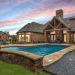 A backyard with a stone-edged swimming pool, lounge chairs, and a garden, set beside a large brick house under a colorful sunset sky.